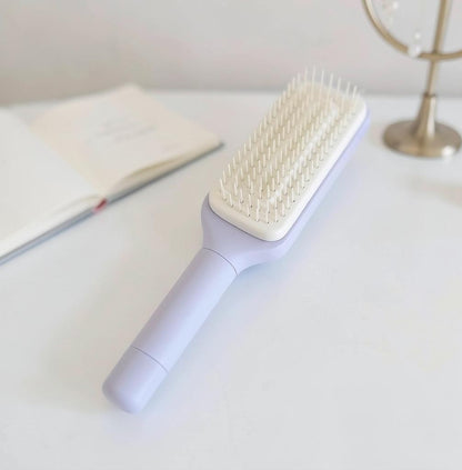 Snaggedly 4-in-1 self-cleaning hair brush in pastel purple with white bristles on a white desk surface.